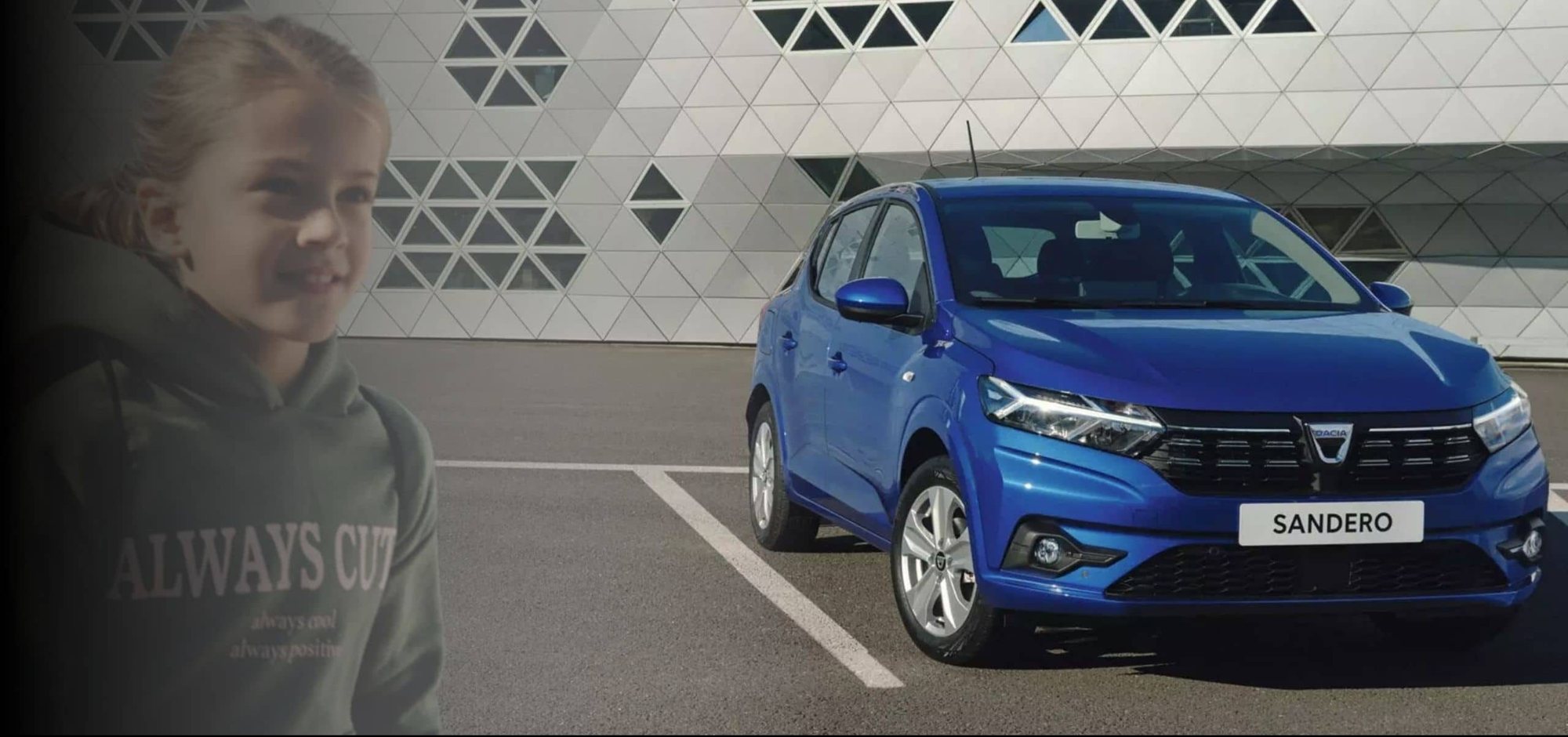 Une Dacia Sandero bleue est garée sur un parking devant un bâtiment à façade géométrique, à côté d'une fillette en sweat à capuche vert.