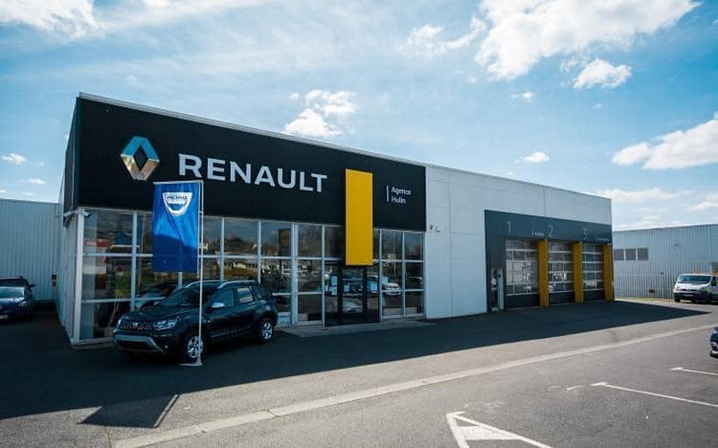 Façade vitrée d'une agence Renault sous un ciel bleu, avec un SUV Dacia noir garé devant et des portes d'atelier à droite.