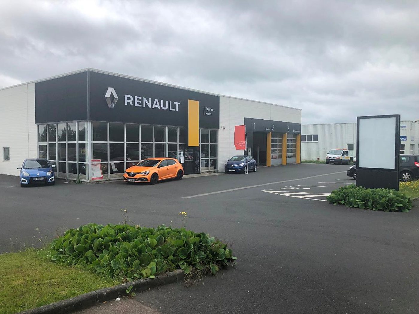 Façade d'une agence Renault avec une Mégane orange et deux autres voitures garées devant, sous un ciel nuageux.