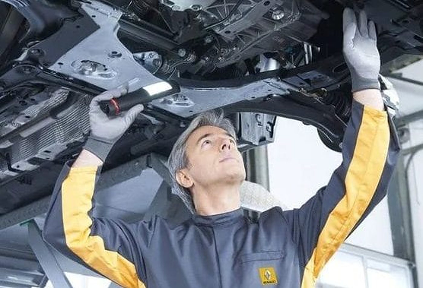 Un mécanicien en combinaison grise et jaune, portant des gants, éclaire avec une lampe le dessous d'une voiture surélevée dans un atelier.