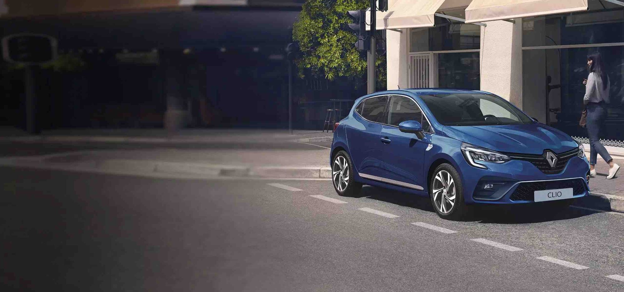 Une Renault Clio bleue est garée au bord d'une rue de ville, tandis qu'une femme marche sur le trottoir près d'une vitrine.