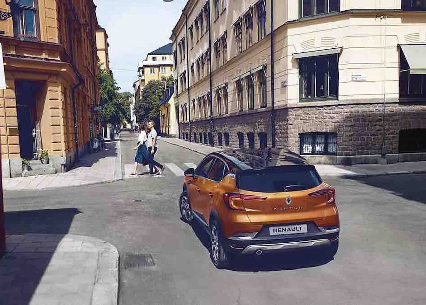 Un SUV Renault orange vu de l'arrière tourne à un carrefour d'une rue bordée d'immeubles anciens, pendant qu'un couple traverse sur le passage piéton.