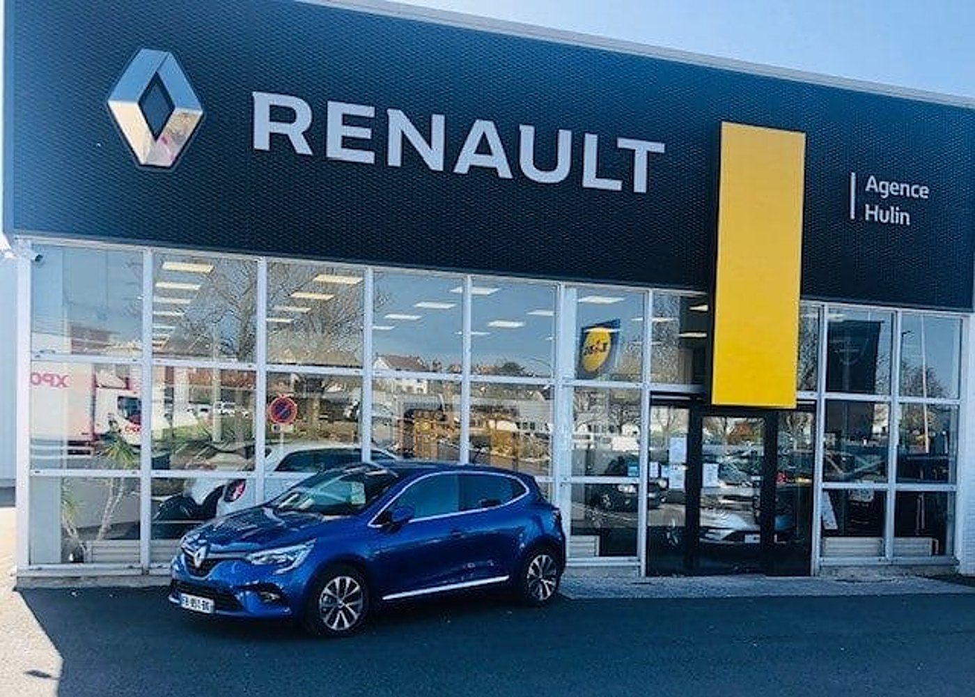 Une Renault Clio bleue est garée devant la façade vitrée de l'agence Renault Hulin, sous une enseigne noire et jaune.